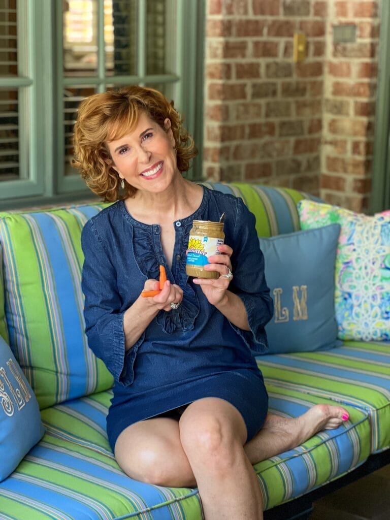 woman wearing blue dress holding sunbutter and carrots sitting on a colorful sofa