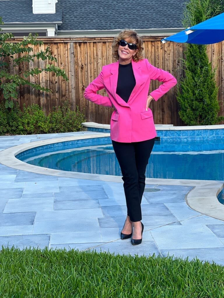 woman in sunglasses standing by a pool wearing black pants and pumps with a hot pink blazer