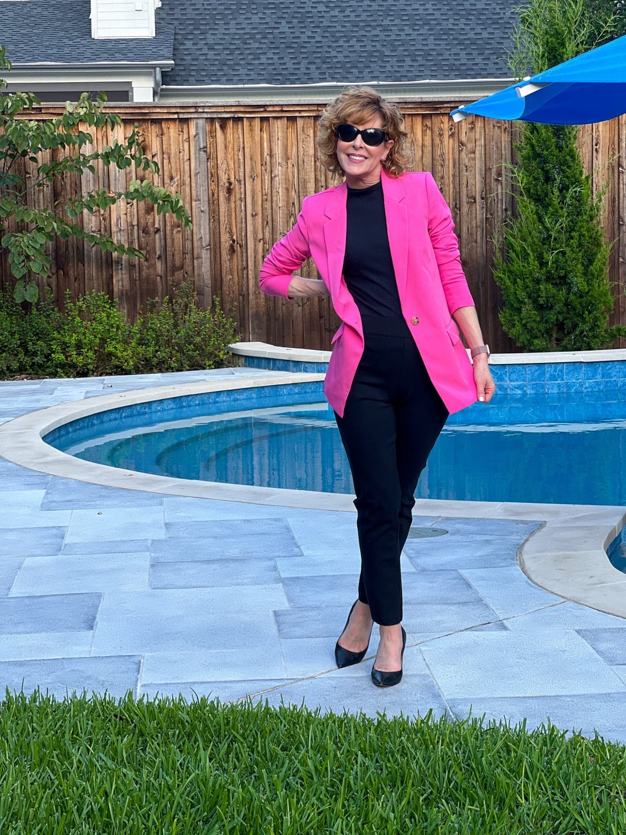 woman in sunglasses standing by a pool wearing black pants and pumps with a hot pink blazer unbuttoned
