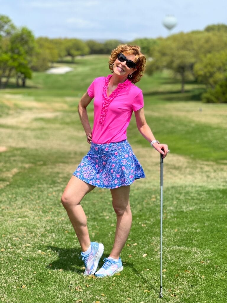 woman wearing sunglasses standing on a golf course holding a golf club wearing lilly pulitzer frida polo top and coordinating skort