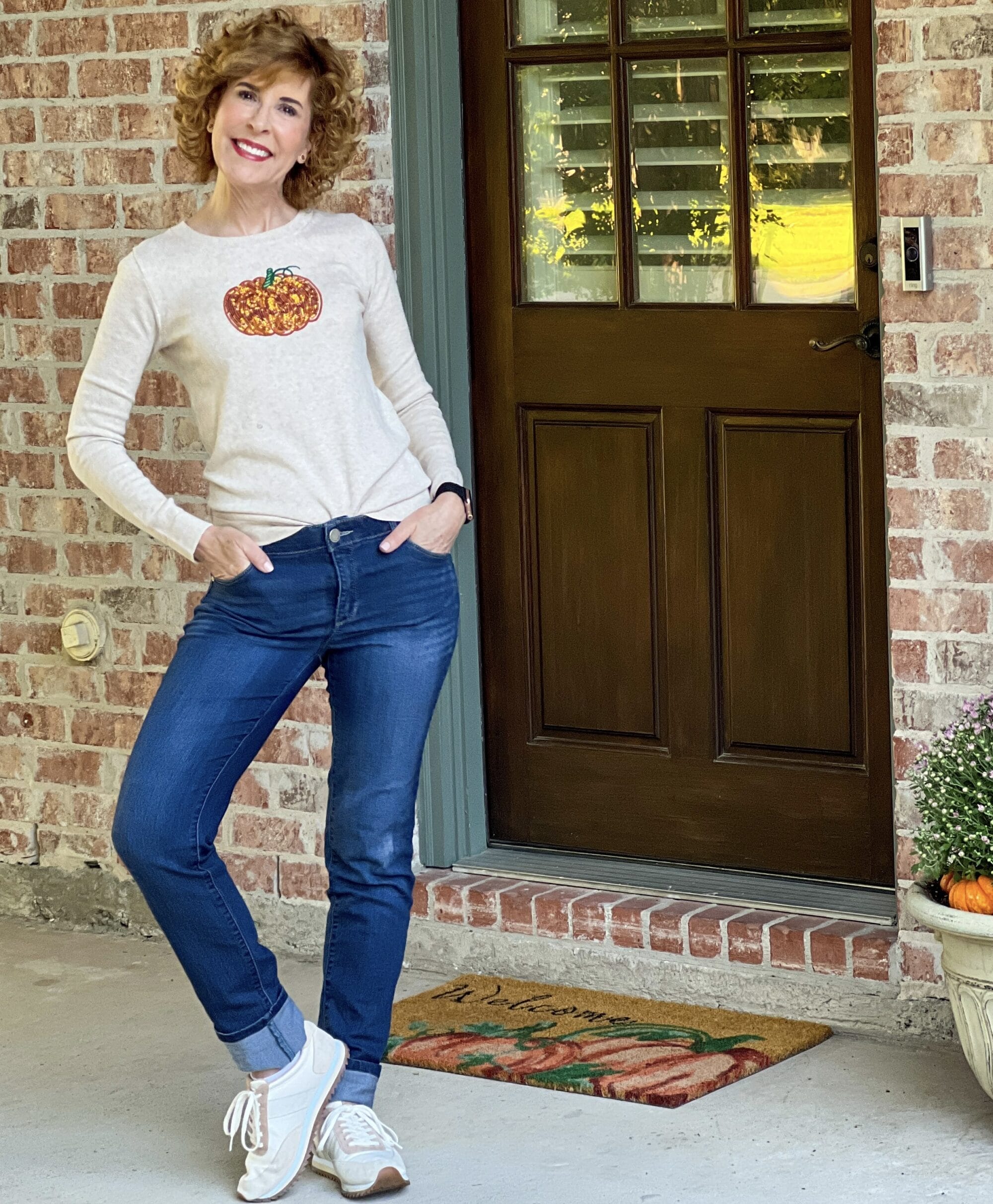 woman over 50 standing on her back porch wearing a j.Crew factory sequin pumpkin sweater and jeans with j.crew tan sneakers