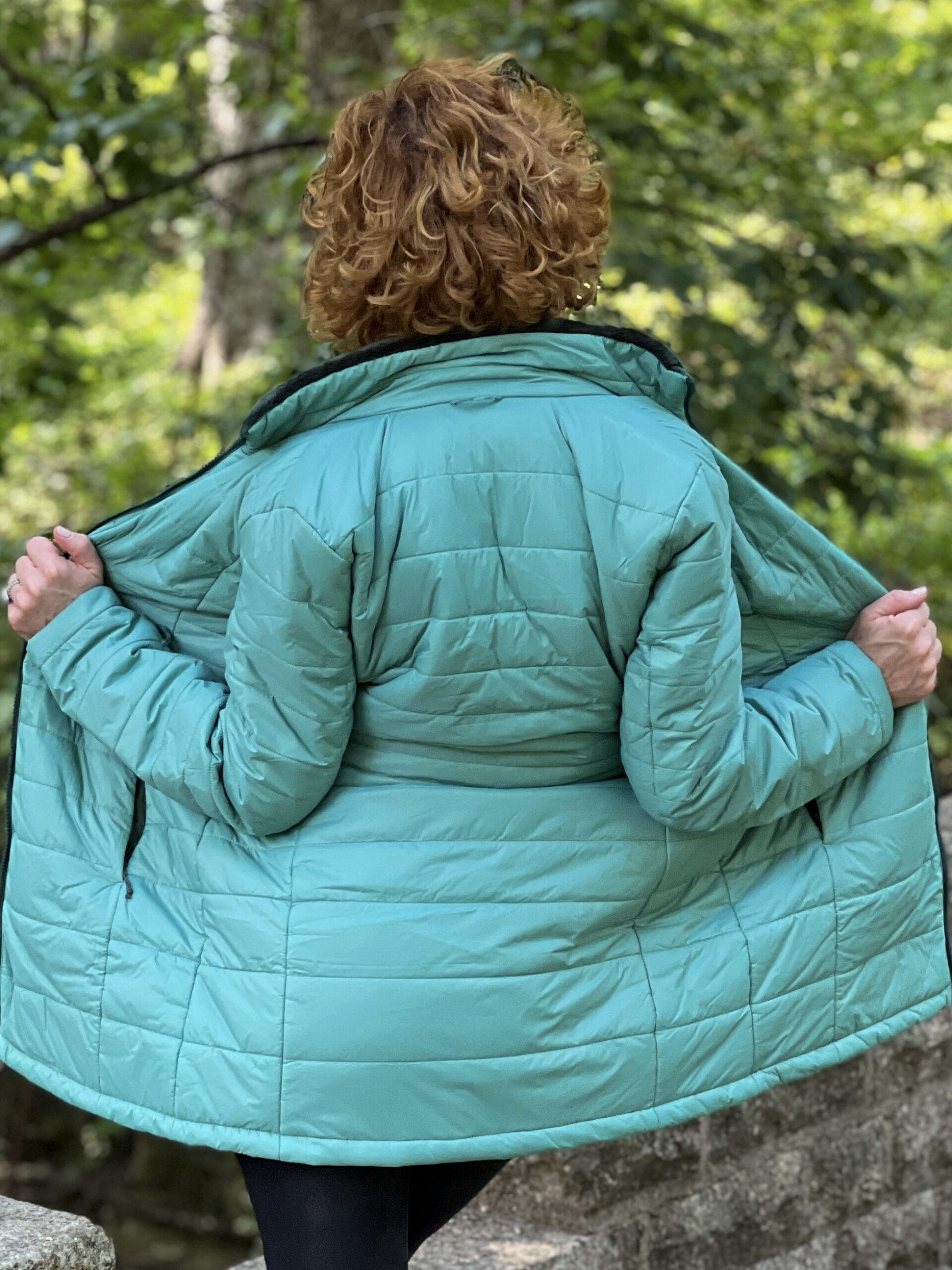 woman with brown curly hair holding open the green primaloft puffer jacket from the back