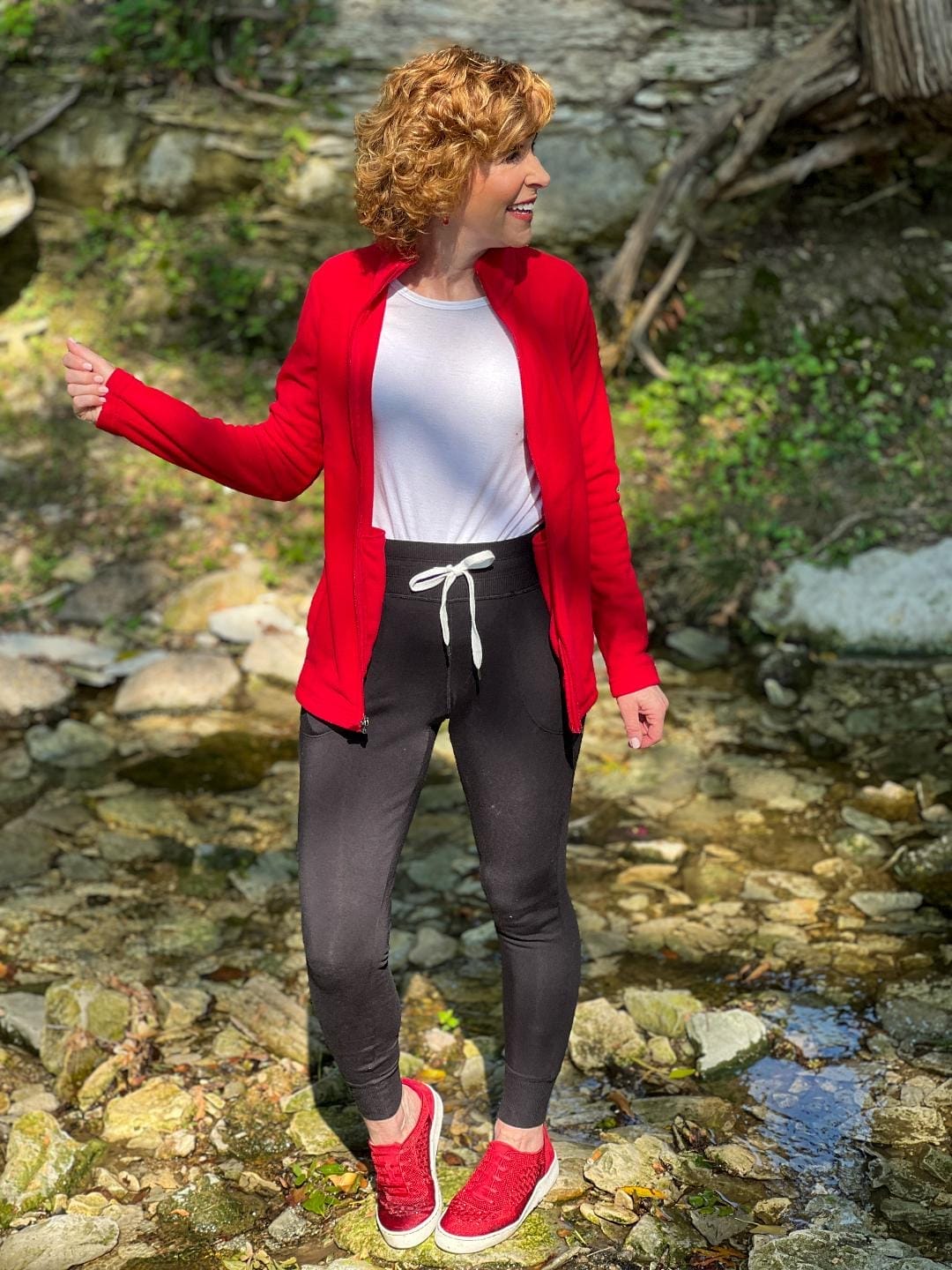 woman wearing lands' end red fleece a white tee and black joggers with red shoes stepping on rocks while crossing a creek bed