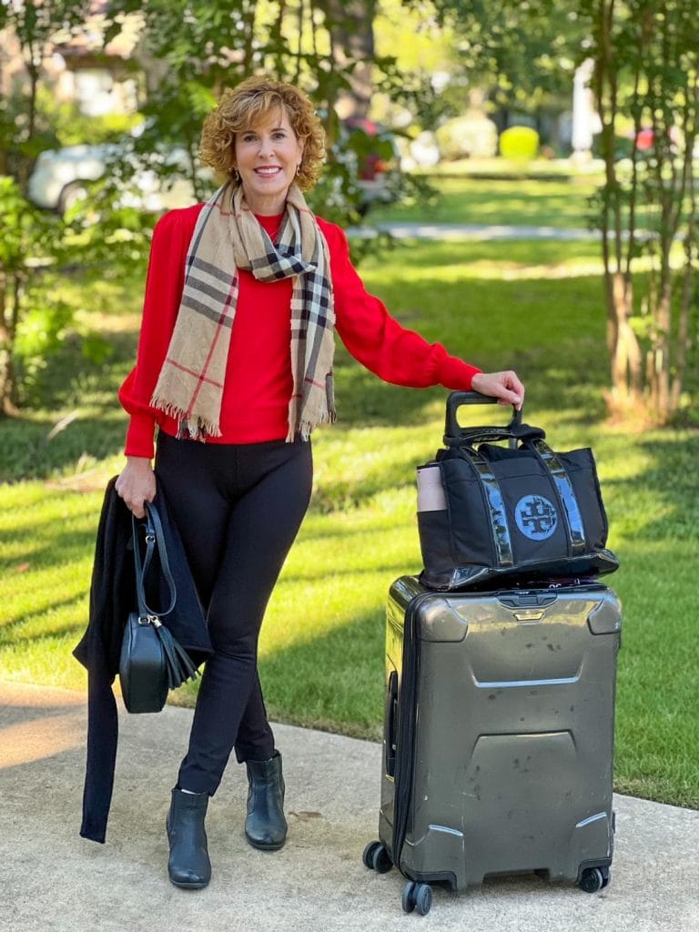 woman over 50 standing on driveway going on Nyc trip wearing spanx black perfect pants, black booties, red cashmere sweater, burberry scarf with suitcase and tory burch tote