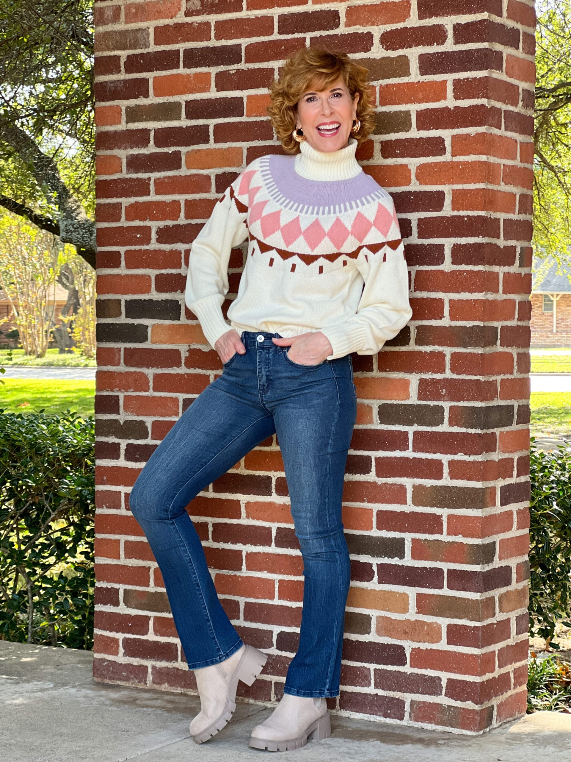 Woman over 50 standing by a brick wall wearing Free Assembly Women's Fair Isle Sweater, Sofia Jeans by Sofia Vergara Women's High Rise Skinny Kick Bootcut Jeans, and Madden NYC Women's Lug Sole Chelsea Booties all from Walmart
