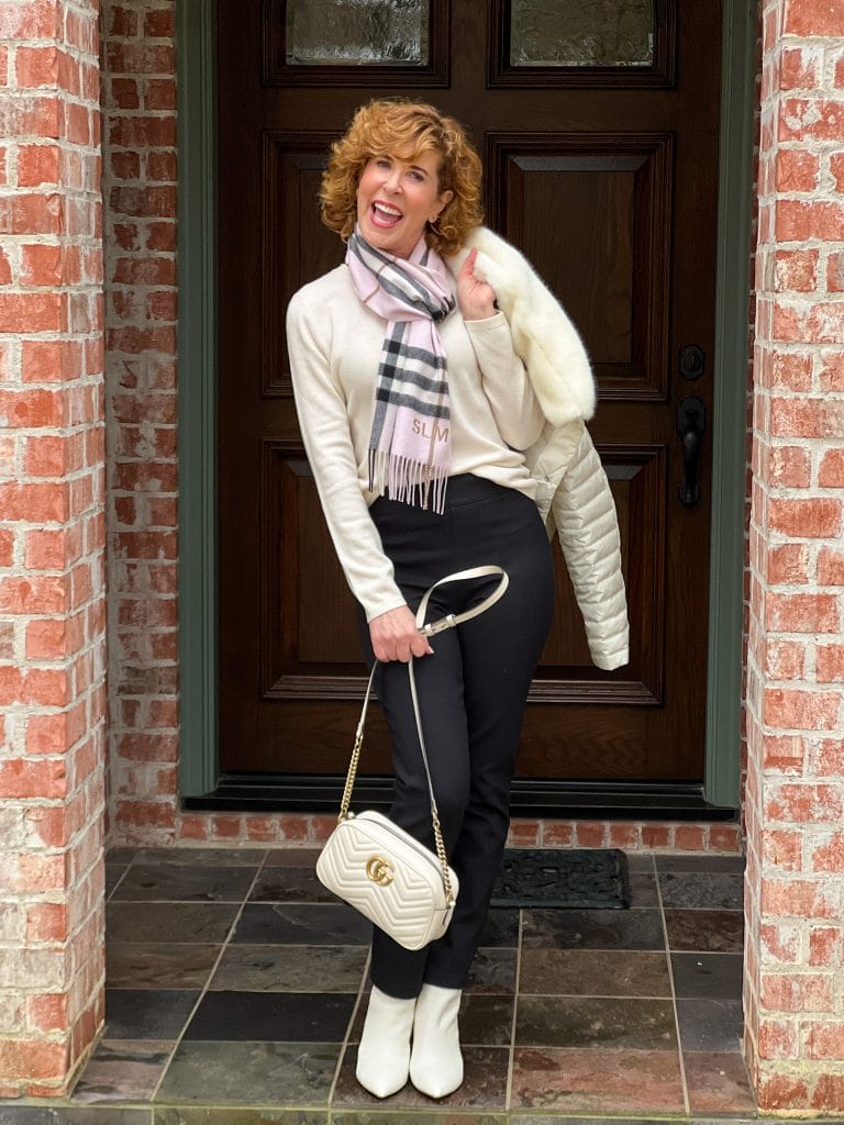 woman over 50 standing on front porch wearing ivory cashmere sweater with burberry scarf and black spanx perfect ankle pants with black suede booties carrying an ivory gucci handbag and wearing ivory booties