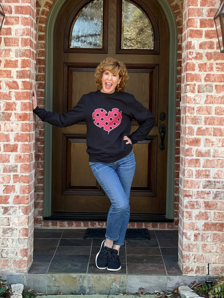 woman over 50 standing on front porch wearing black sneakers, jeans, and a black sweatshirt with viva magenta polka dot heart on it