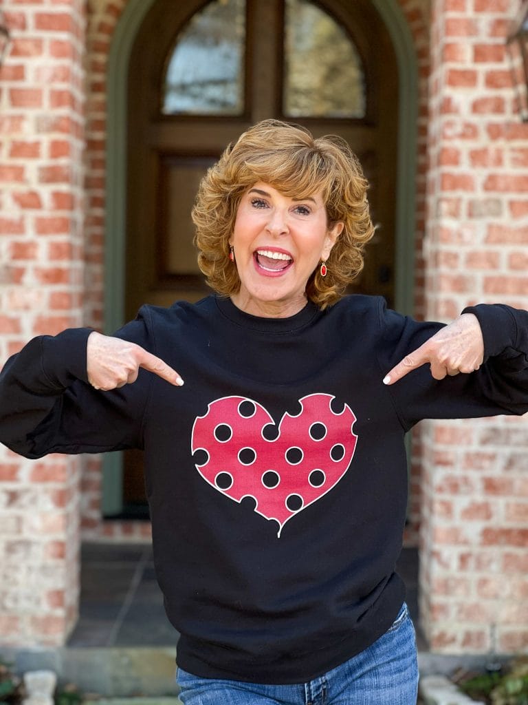 woman over 50 standing in front of front porch pointing to a black sweatshirt with viva magenta polka dot heart on it that she is wearing