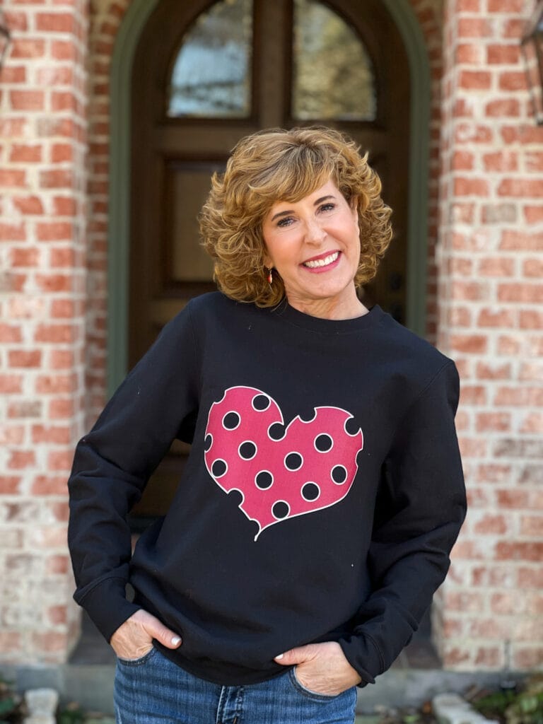 woman over 50 standing in front of front porch wearing a black sweatshirt with viva magenta polka dot heart on it