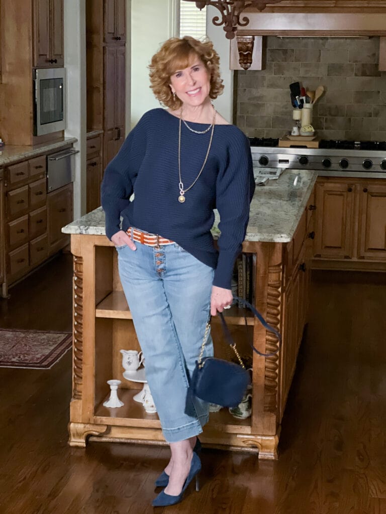 woman over 50 standing in a kitchen wearing cabi's spring 2023 palm beach crop jeans, utopia pullover, utility belt, timepiece earrings, timepiece necklace and navy blue pumps holding a navy blue handbag