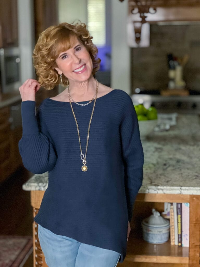 woman over 50 standing in a kitchen wearing cabi's spring 2023 utopia pullover, utility belt, timepiece earrings, timepiece necklace