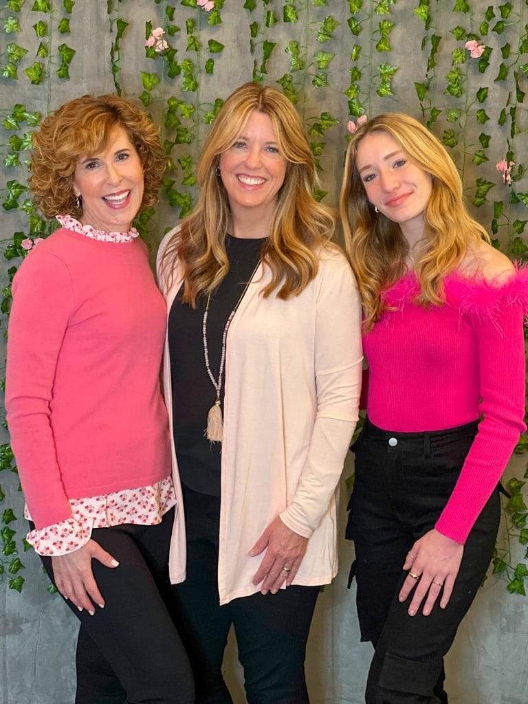 three women dressed in shades of pink in front of an ivy & flower wall backdrop