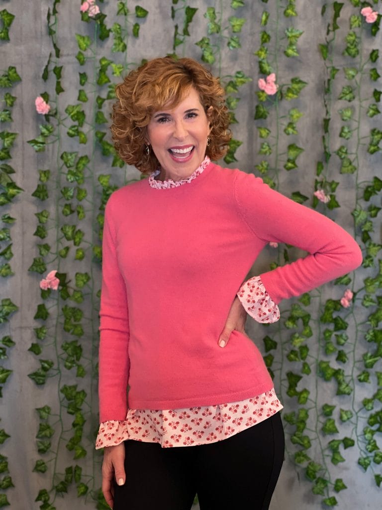 woman over 50 wearing hill house home pink ruffle neck top and coral pink cashmere sweater standing in front of an ivy and pink flower photo backdrop