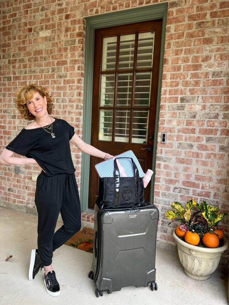 woman over 50 wearing black amazon jumpsuit and black sneakers standing by luggage