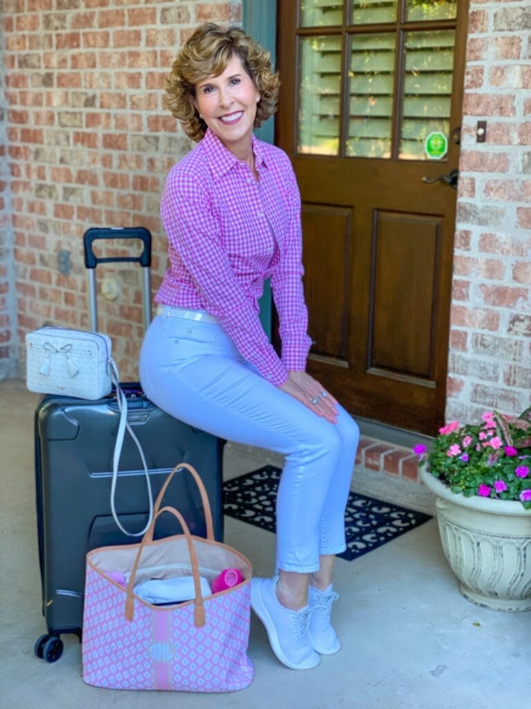 woman over 50 sitting on suitcase wearing pink and white gingham button down and white girlfriend jeans next to luggage