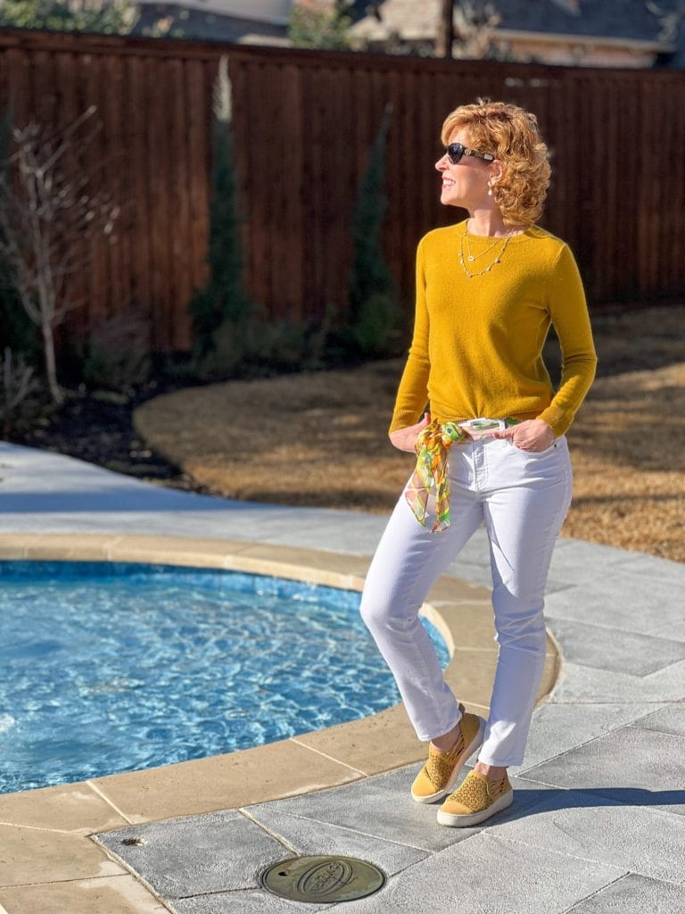 woman standing by a pool wearing yellow cashmere sweater, white jeans, yellow sneakers