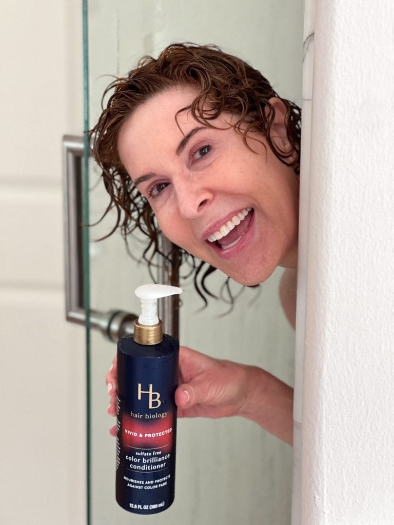 woman peeking out of a shower with shampoo in her hair holding hair biology vivid and protected