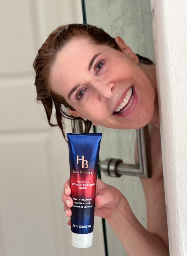 woman peeking out of a shower with shampoo in her hair holding hair biology vivid and protected conditioning mask