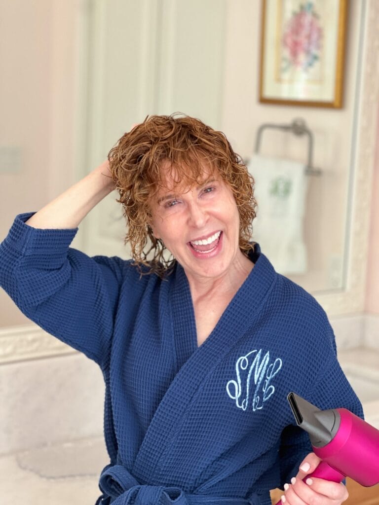 woman over 50 wearing blue bathrobe standing in bathroom with curly wet hair