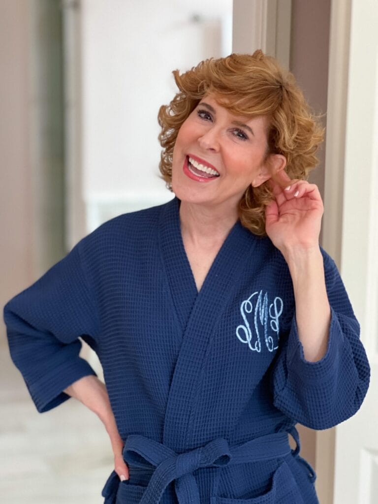 woman over 50 wearing blue monogrammed robe tucking a strand of her brown curly hair behind her left ear