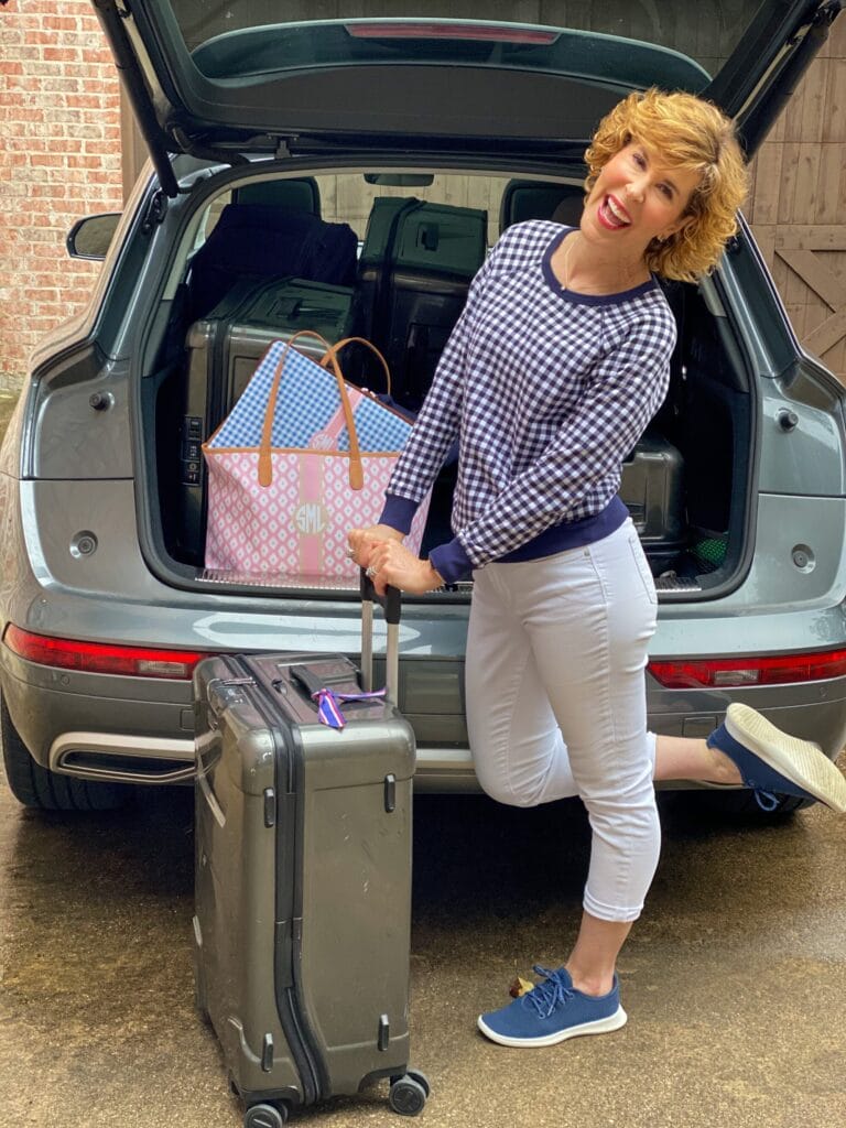 travel look for woman over 50 standing by the open trunk of the car wearing navy and white gingham sweatshirt and white crop jeans with navy sneakers standing by luggage