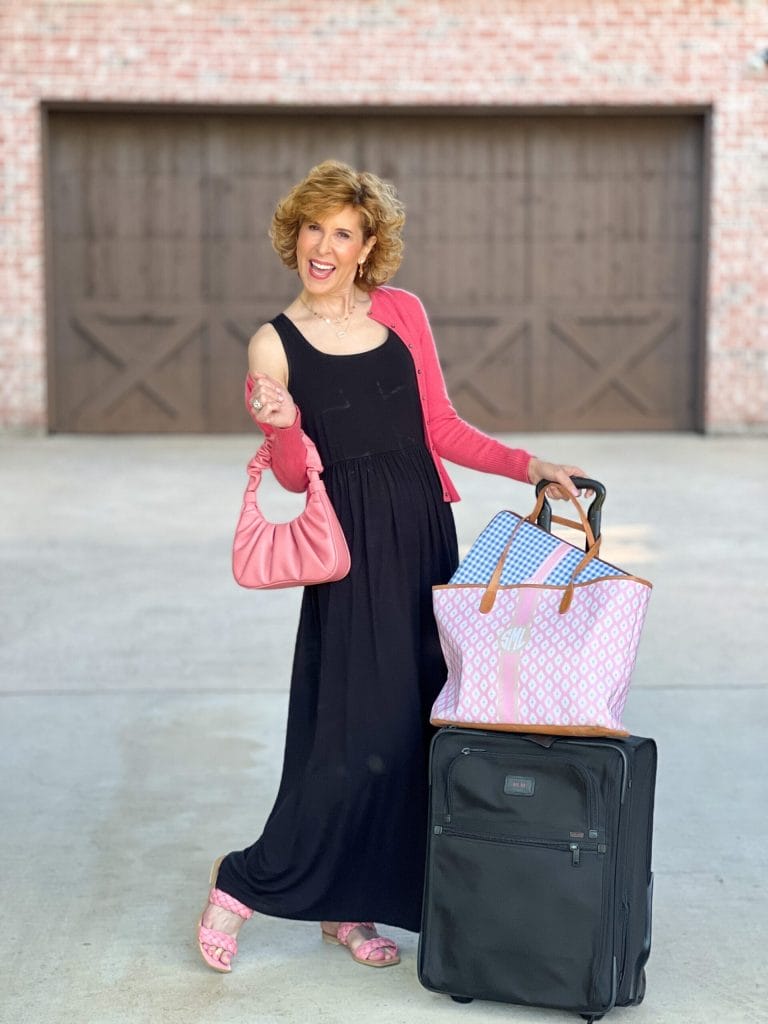Woman over 50 wearing black maxi dress with coral cardigan and purse standing by suitcase and travel totes standing on driveway