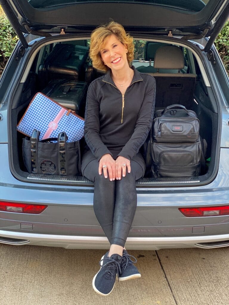 woman over 50 sitting in the trunk of a car wearing faux leather leggings and lilly pulitzer black dress with travel tote and backpack on either side of her