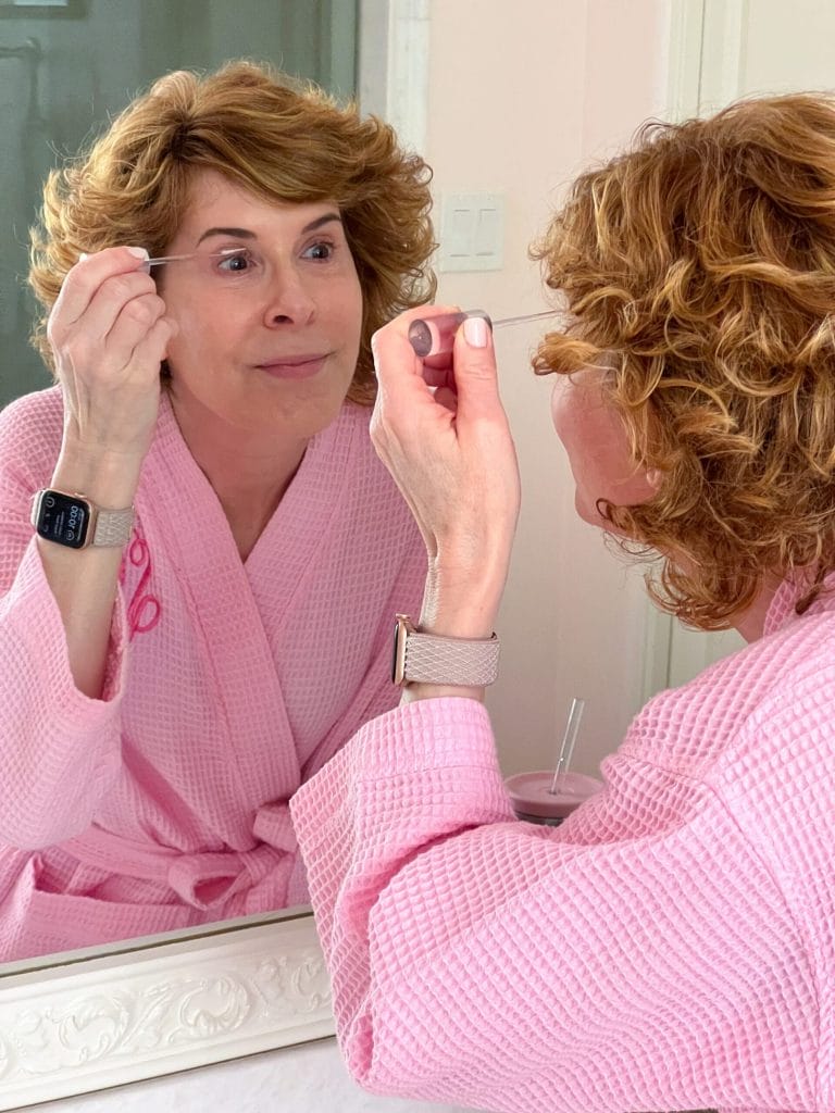 woman over 50 dressed in a pink robe looking into a mirror using an eyelash serum