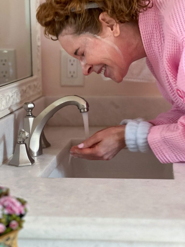 woman over 50 dressed in pink robe rinsing after cleansing face