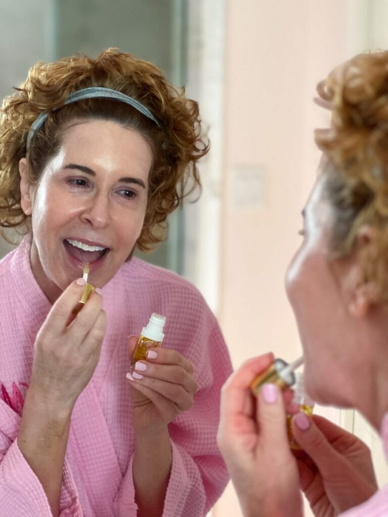 woman over 50 dressed in pink robe applying lip oil as a part of her skincare over 50 nightime regimen