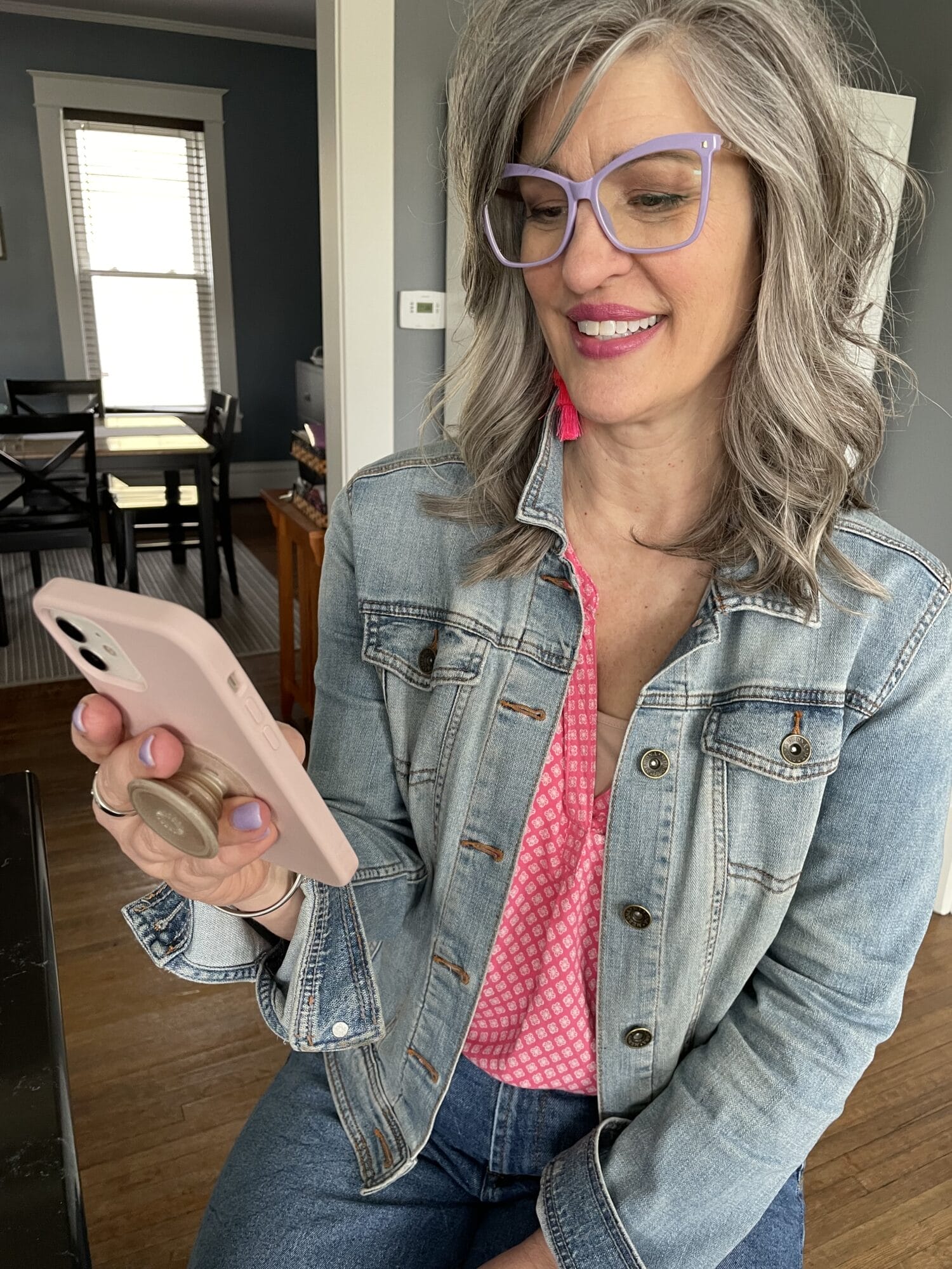woman wearing pink top a denim jacket, and big glasses looking at her cell phone in her house