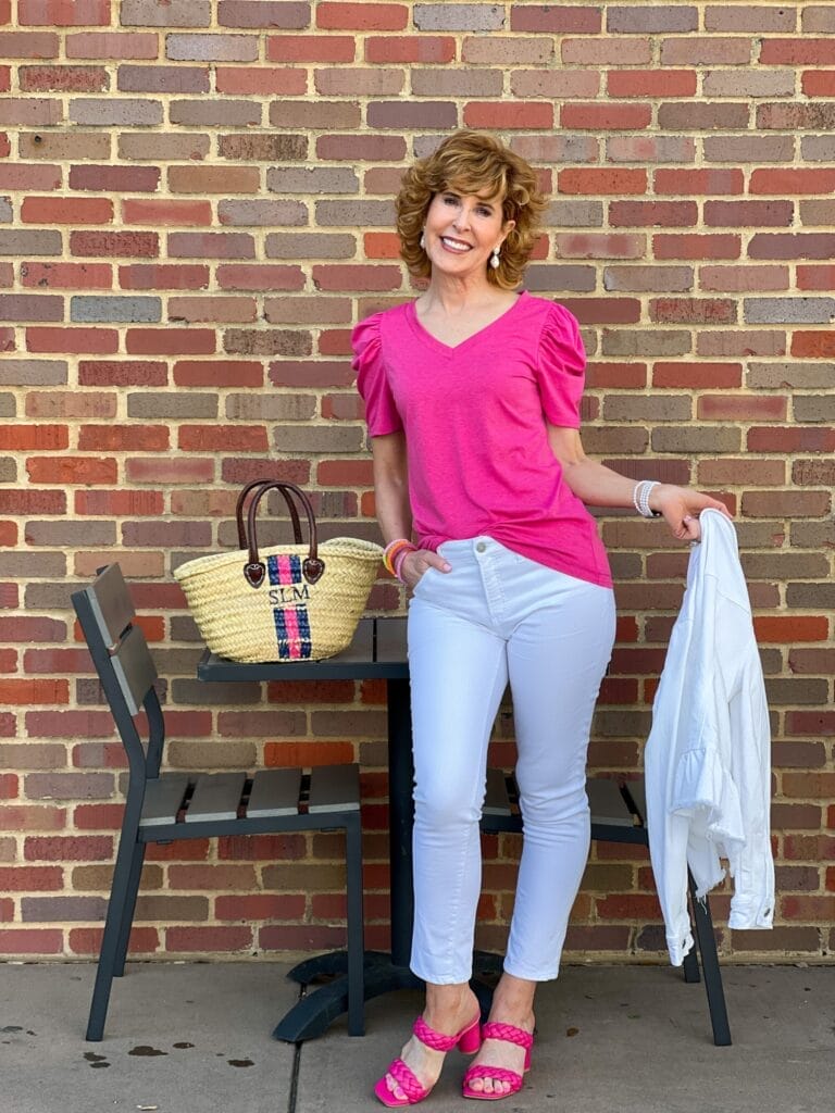 woman holding chico's white peplum denim jacket and wearing white girlfriend jeans with pink amazon v-neck tee and pink target heels standing at a table outdoors next to a handpainted monogrammed straw bag thinking about how to find her purpose in the empty nest