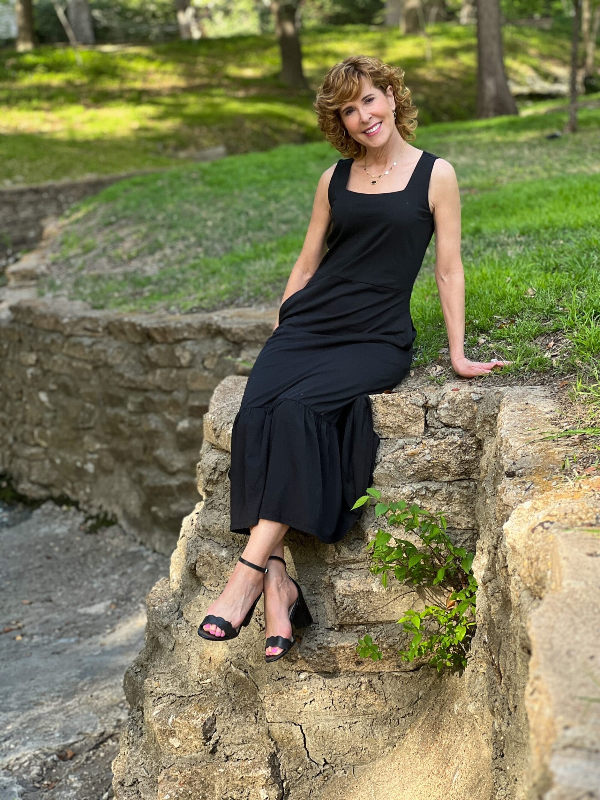 woman sitting on stone wall wearing black tank style dress and black ankle strap sandals