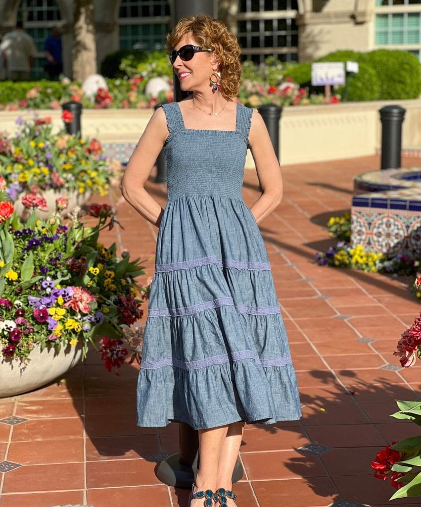 woman wearing sunglasses and a chambray tiered skirt dress from lands' end posing by a container of flowers on a sunny day