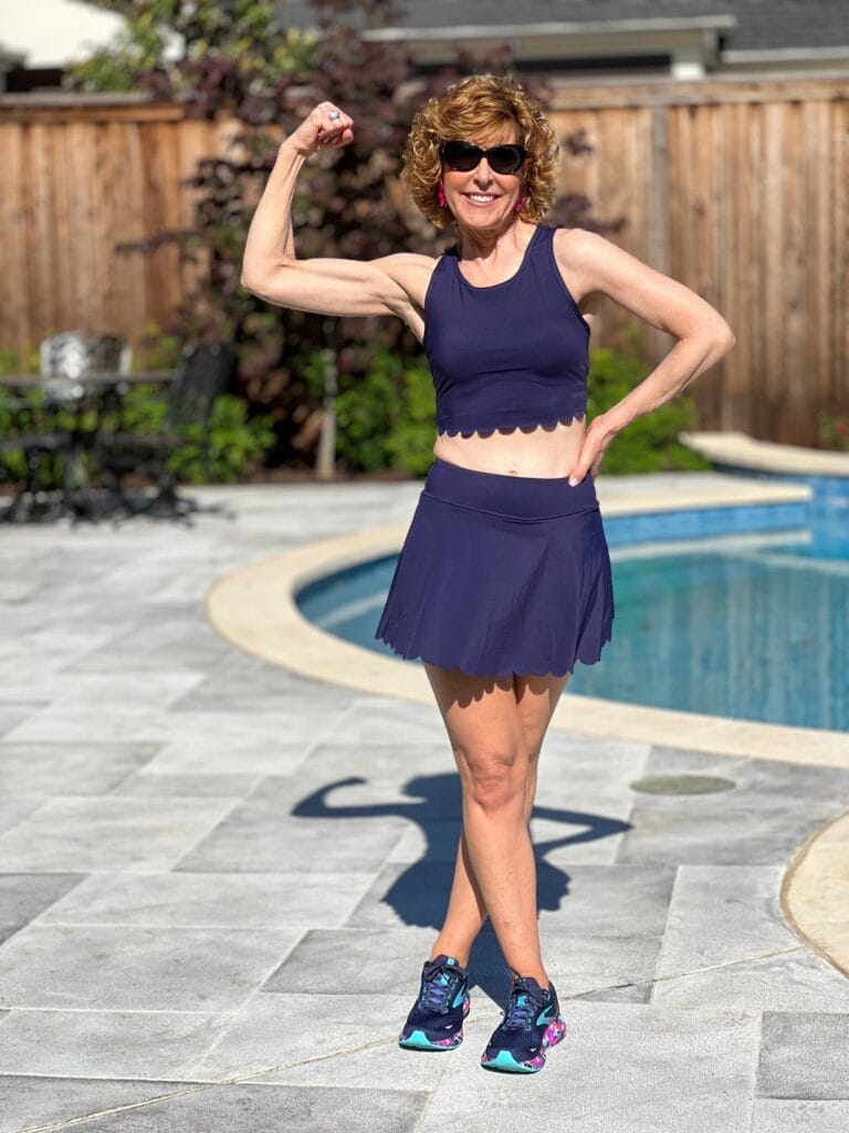 woman over 50 wearing navy blue scalloped workout set standing by a pool making a muscle with her right arm