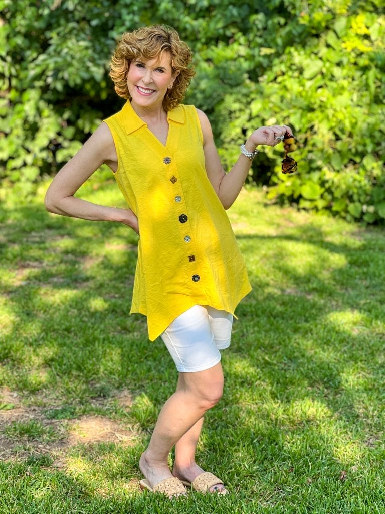 woman over 50 wearing yellow tunic top and white long shorts standing outside