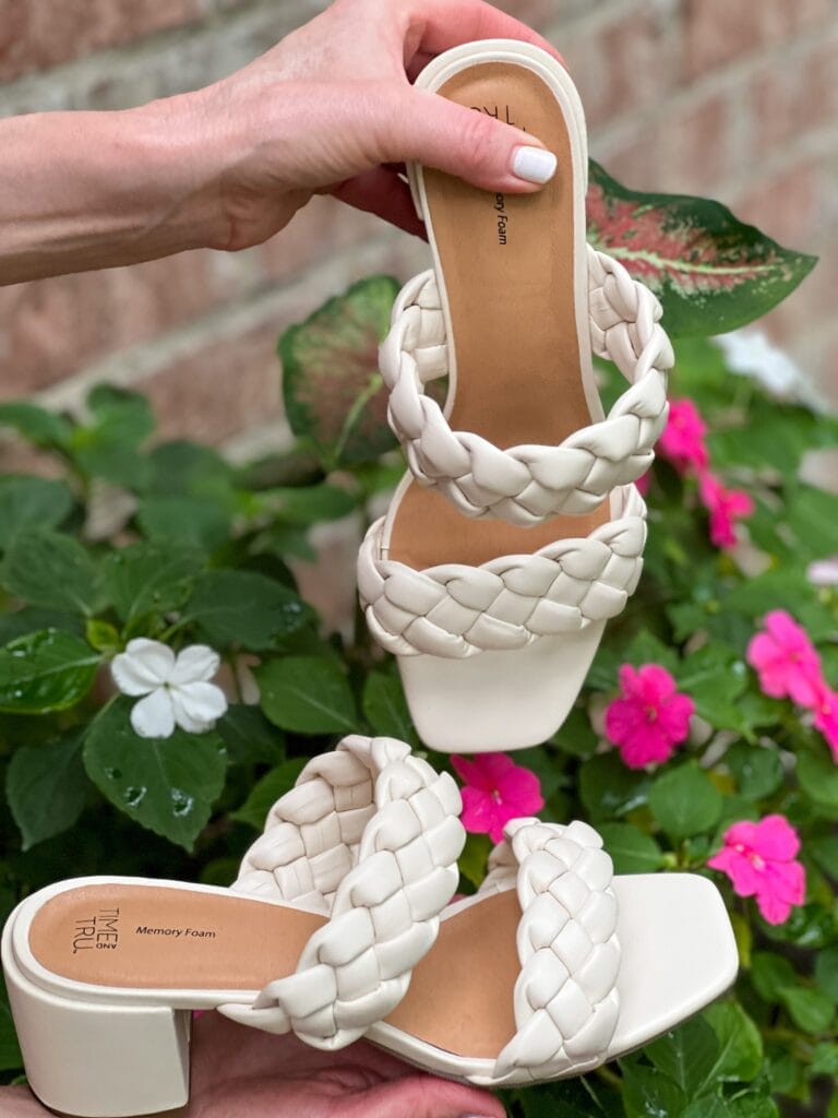 pair of off-white braided two-strap block heel sandals being held by a woman's hands in front of a flower pot of pink and white flowers