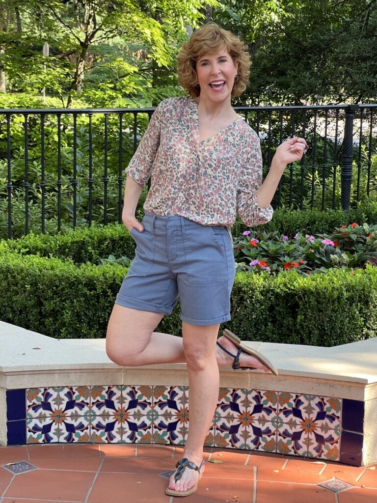 woman standing near a colorful short wall with one leg up on it wearing blue cargo shorts and a pintuck blouse from nydj