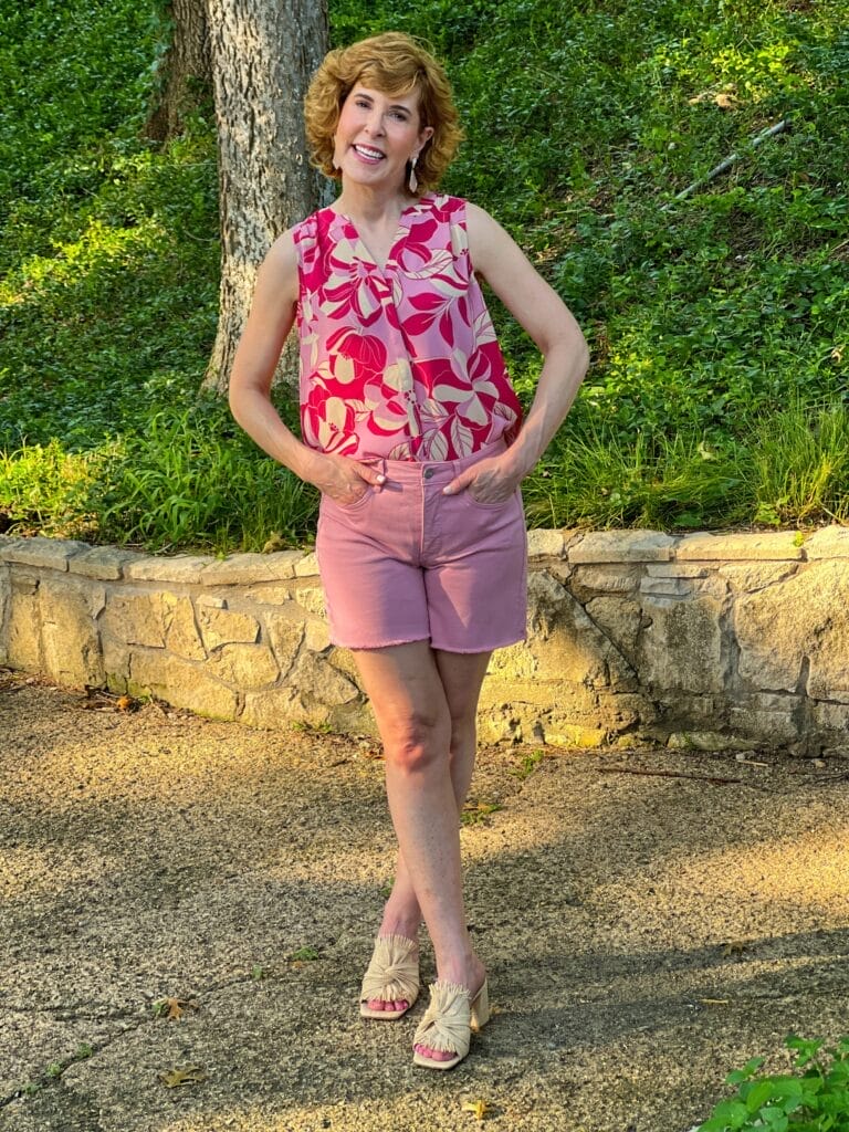 woman over 50 posing on a gravel path wearing nydj a-line denim shorts in pink and sleeveless pintuck blouse in red and pink