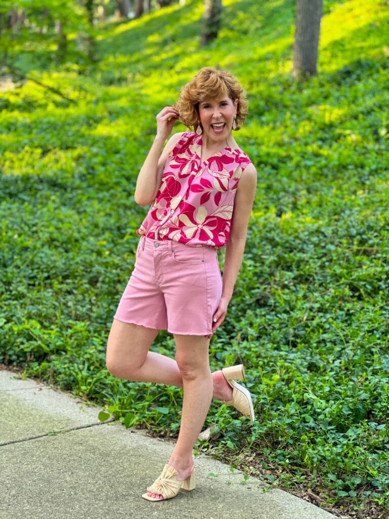 close up of woman over 50 posing wearing nydj a-line denim shorts in pink and untucked sleeveless pintuck blouse in red and pink