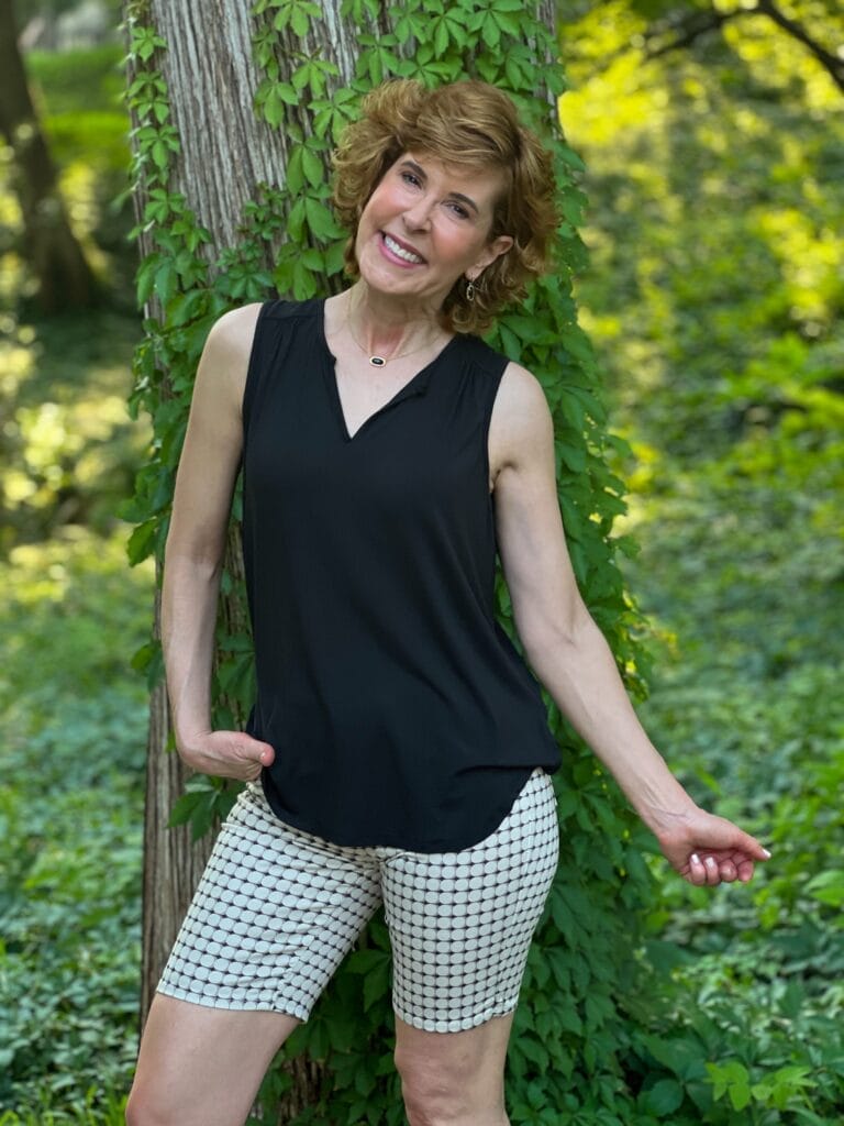 woman over 50 posing beside a vine covered tree wearing a black top and long shorts
