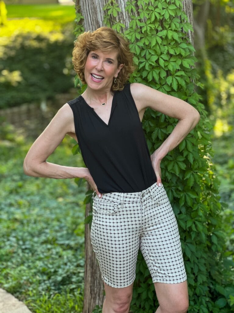 woman over 50 posing with hands on hips wearing black sleeveless tee tucked into long ella shorts by nydj