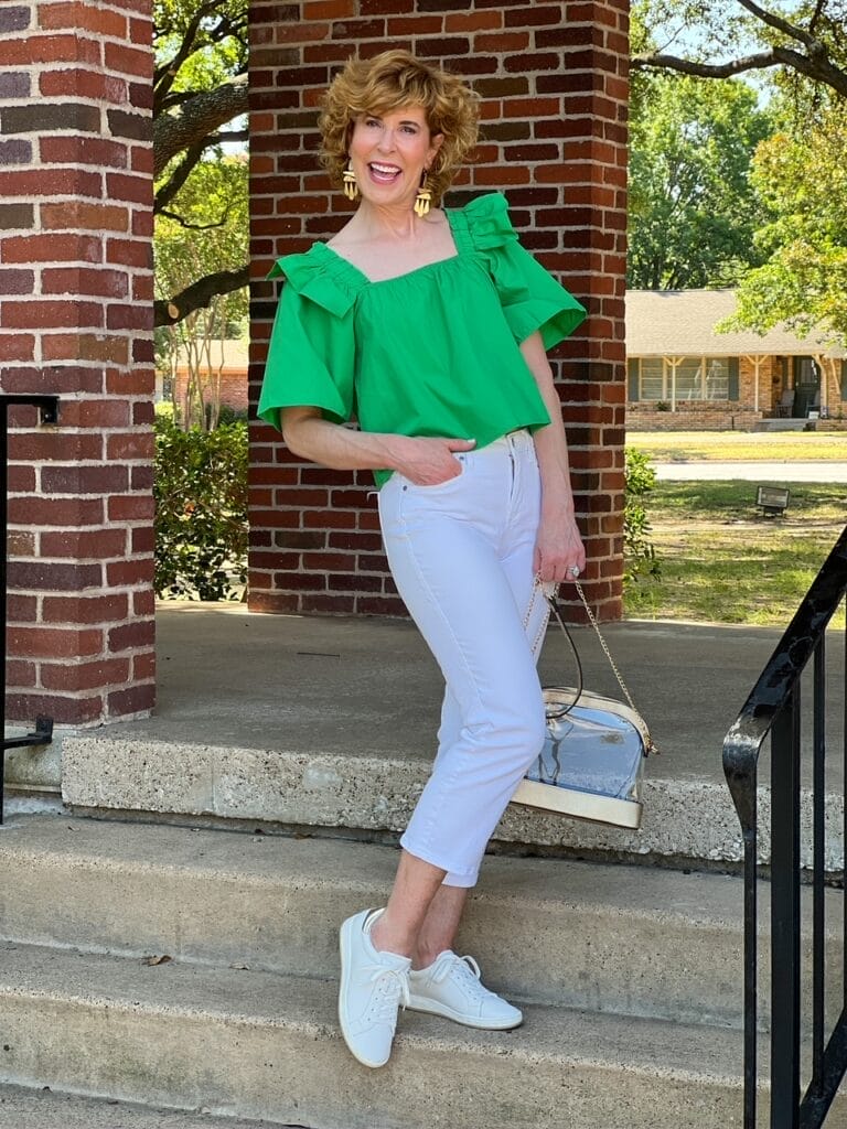 woman in green crop top and white capri jeans and white sneakers standing on steps holding a clear stadium purse