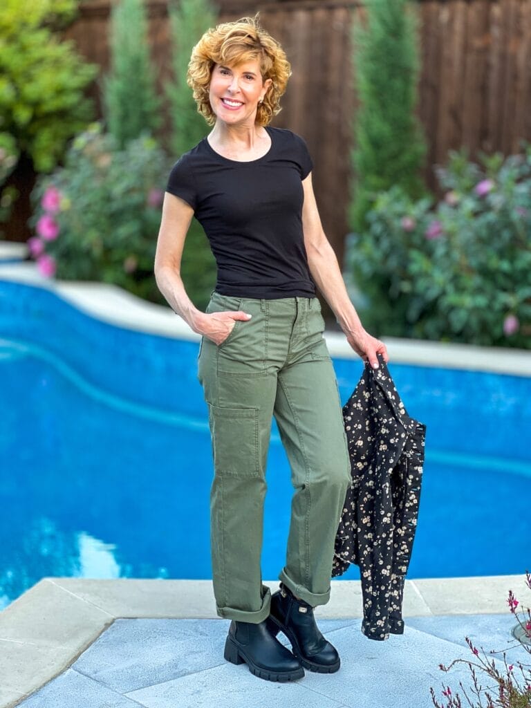 woman over 50 standing by a pool wearing cargo pants and a black tee holding a black floral jacket