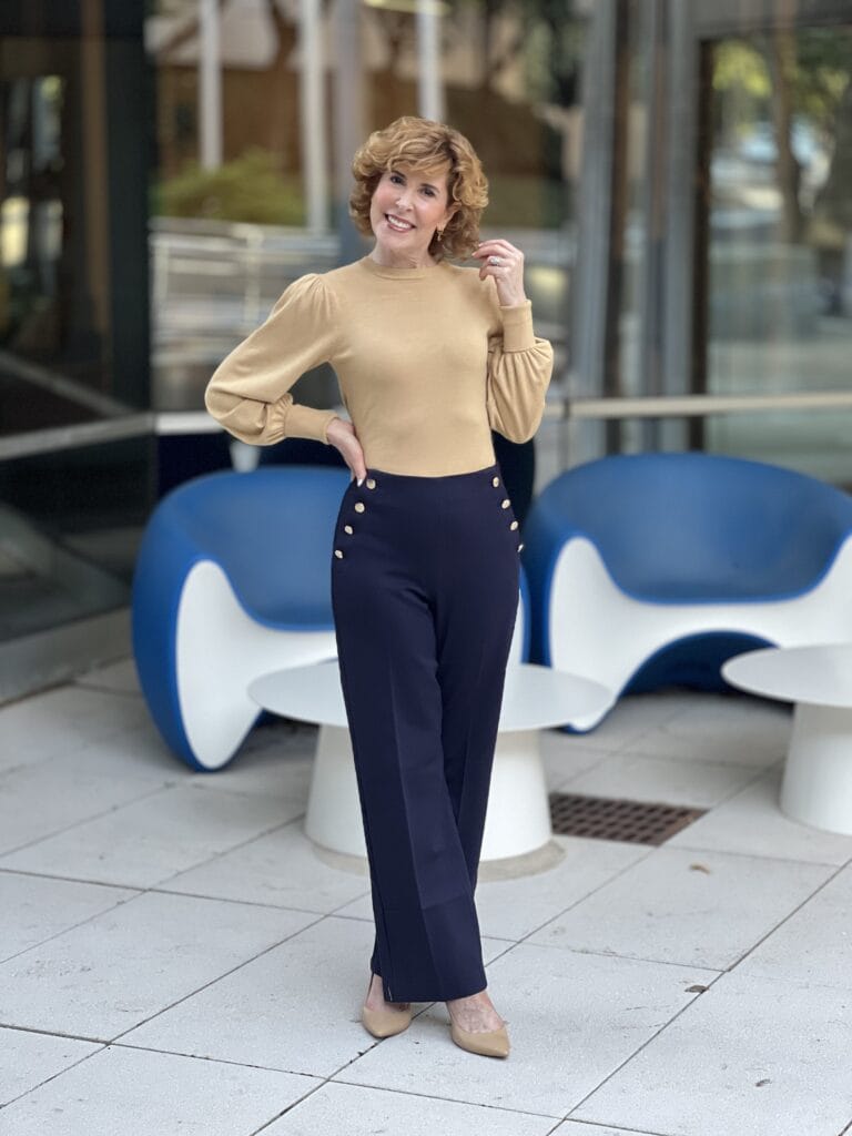 woman touching her hair wearing greenwich pants from talbots with gold amazon sweater standing by blue chairs