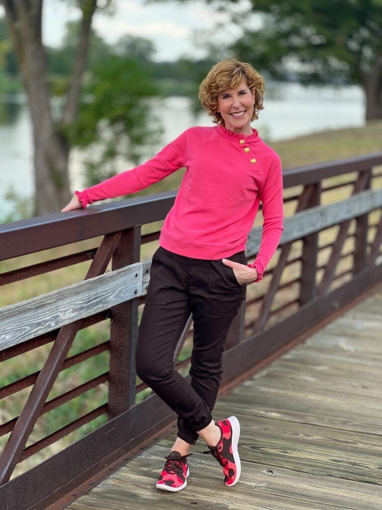 woman on bridge wearing ruffle neck coral sweatshirt with brass buttons and brown joggers with fun sneakers