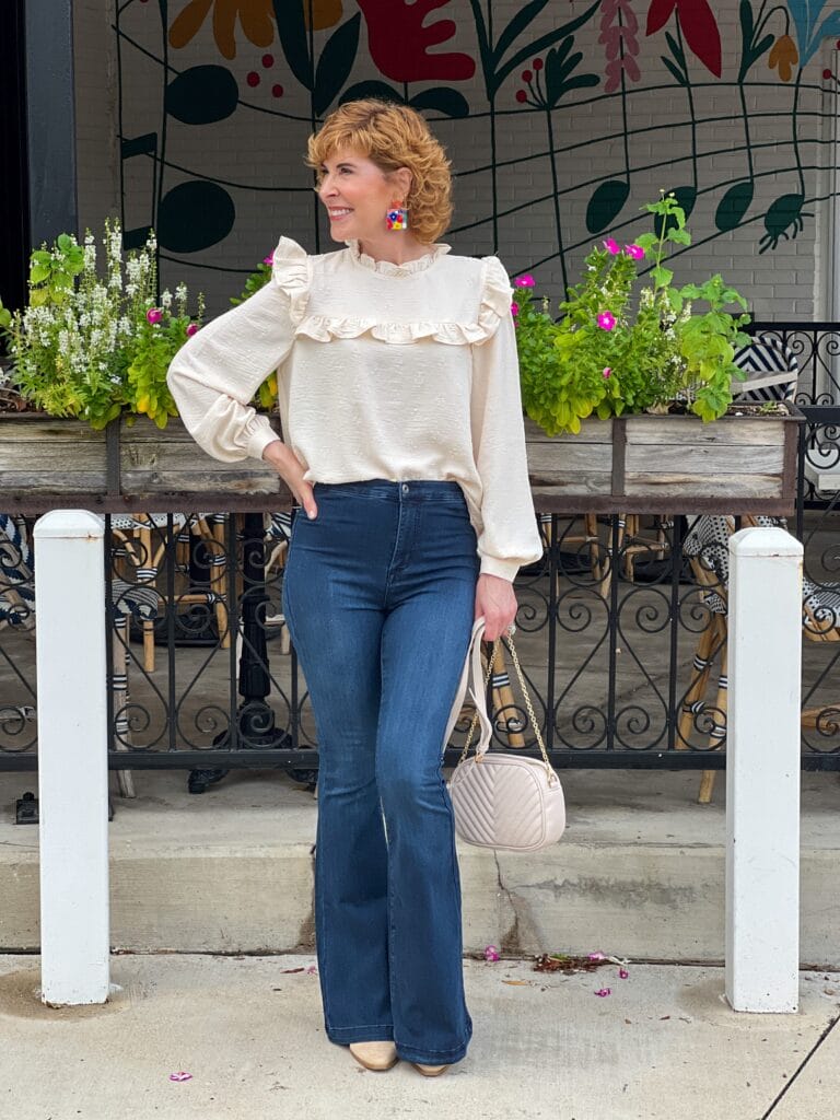 woman over 50 standing outside in front of a restaurant wearing cream ruffled yoke neck blouse, cream quilted crossbody bag, flare jeans, light taupe booties