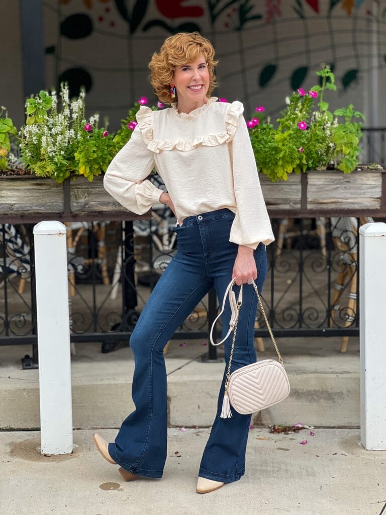 woman over 50 standing outside in front of a restaurant wearing cream ruffled yoke neck blouse, cream quilted crossbody bag, flare jeans, light taupe booties