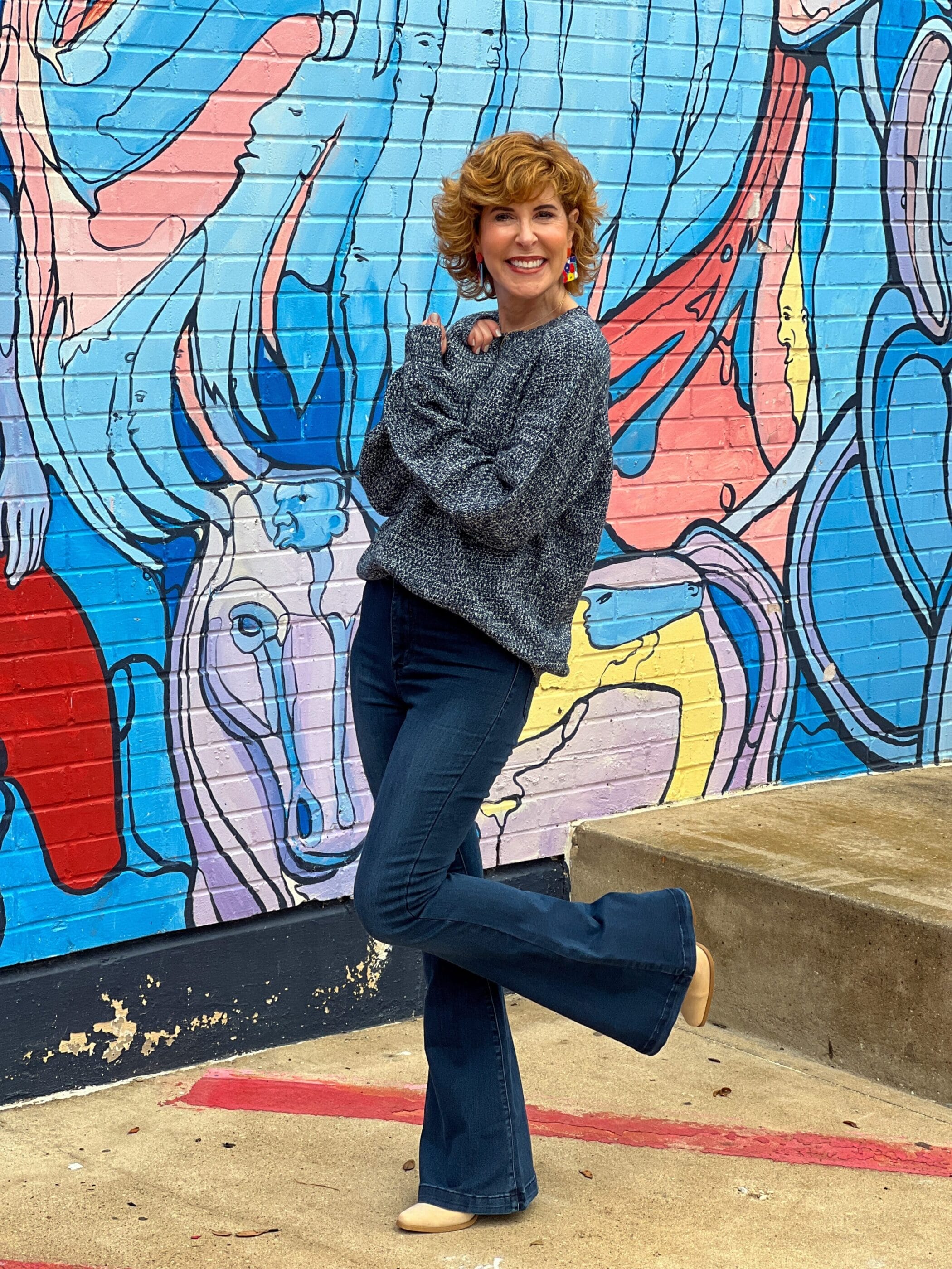 woman wearing flare jeans and a teal sweater with tan booties standing in front of a mural