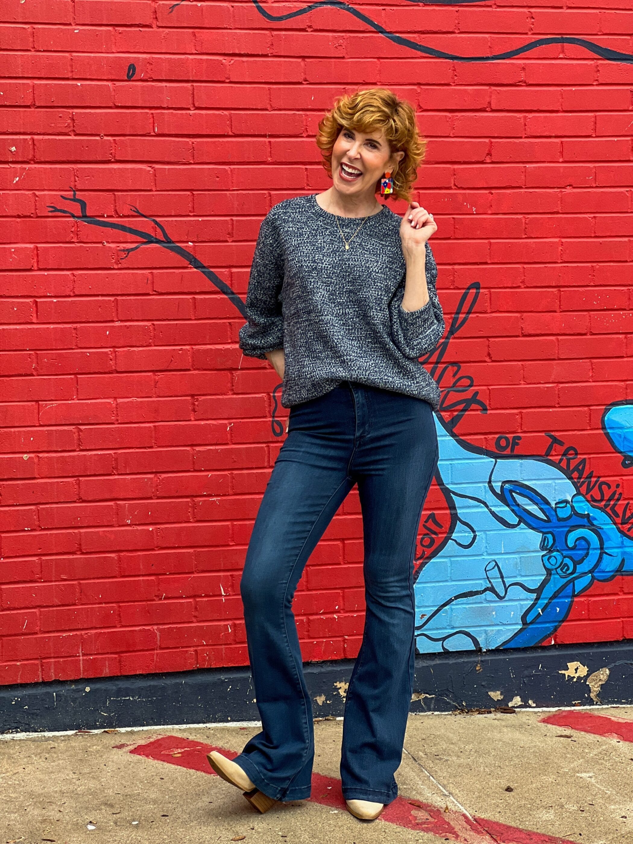 woman over 50 standing in front of a mural wearing teal bubble sleeve sweater and flare jeans from shop the mint